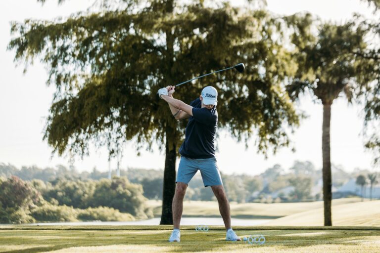 Golfer in mid-swing on a sunny golf course surrounded by trees and open landscape.